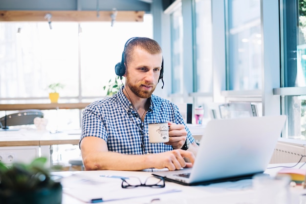 Hombre con taza y headsetworking en la computadora portátil