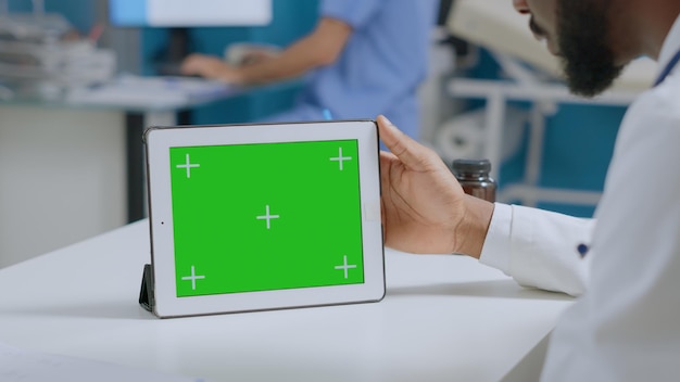 Hombre terapeuta afroamericano sosteniendo simulacros de tableta chroma key de pantalla verde con pantalla aislada en posición horizontal mientras analiza la experiencia en enfermedades en la oficina del hospital. Concepto de medicina