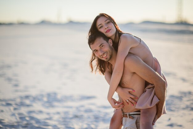 El hombre tiene a una mujer bonita en su espalda caminando en la playa