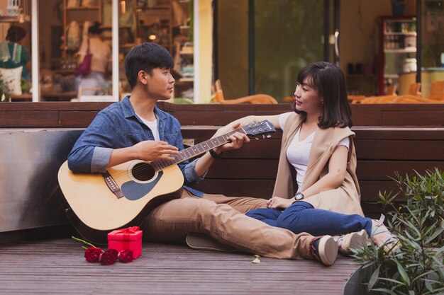 Hombre tocando una guitarra mientras mira a su novia con un regalo y una flor