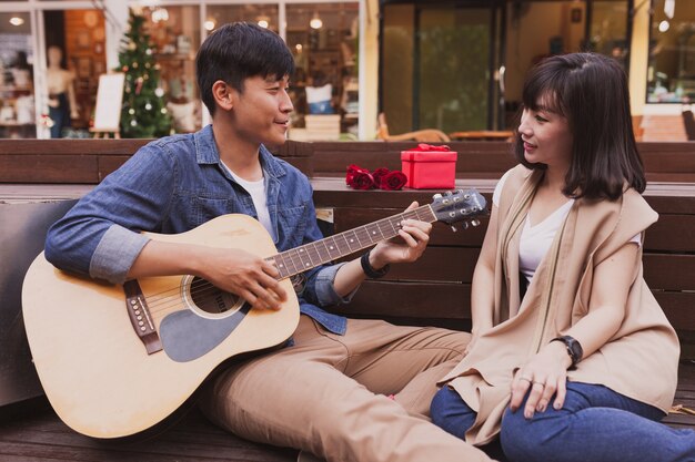 Hombre tocando una guitarra mientras mira a su novia con un regalo y una flor