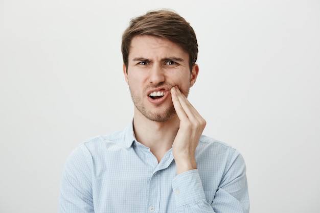 Hombre tocando la mejilla y haciendo muecas de dolor de muelas, necesita dentista