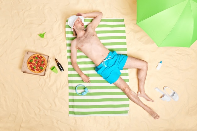 El hombre toma el sol solo y toma la siesta en la playa de arena viste pantalones cortos de Panamá blancos se encuentra en una toalla de rayas verdes descansa junto al mar