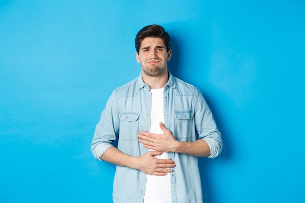 Hombre tomados de la mano en el vientre y haciendo muecas de dolor, quejándose de dolor de estómago, de pie contra el fondo azul.