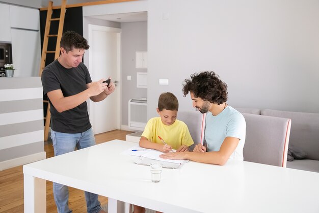 Hombre tomando foto de lindo hijo y esposo haciendo tareas escolares en casa, escribiendo o dibujando en papeles. Concepto de familia y padres gay