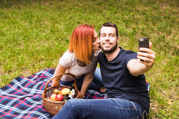 Foto gratuita hombre tomando selfie a través del teléfono móvil con su novia besándolo en la mejilla en un picnic