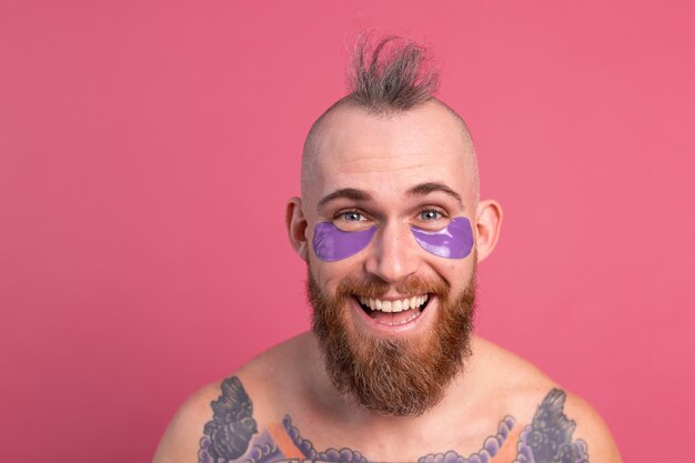 Hombre en topless tatuado barbudo guapo europeo con máscara de parches de ojos morados posando para la cámara en rosa