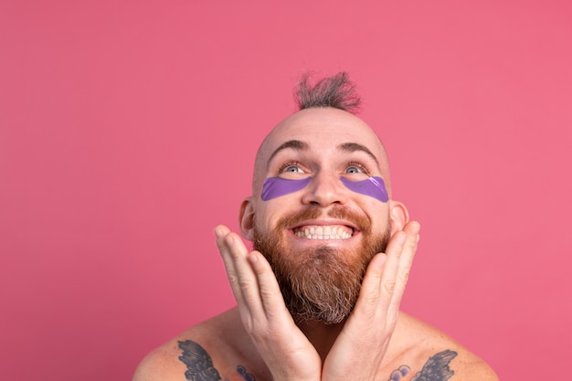 Hombre en topless tatuado barbudo guapo europeo con máscara de parches de ojos morados posando para la cámara en rosa