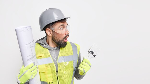 El hombre trabajador industrial posa con planos y pincel de pintura lleva casco, gafas protectoras, uniforme