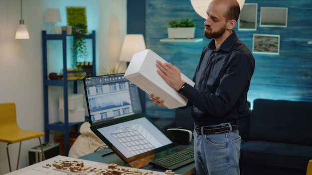 Hombre trabajando como arquitecto analizando maquetas en la mano. Joven ingeniero revisando el modelo de construcción con el plan de planos en el monitor de la computadora para el diseño de la construcción y el proyecto de desarrollo