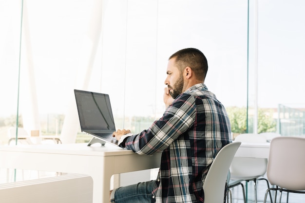 Hombre trabajando frente a la computadora portátil y hablando con el móvil en un espacio de trabajo
