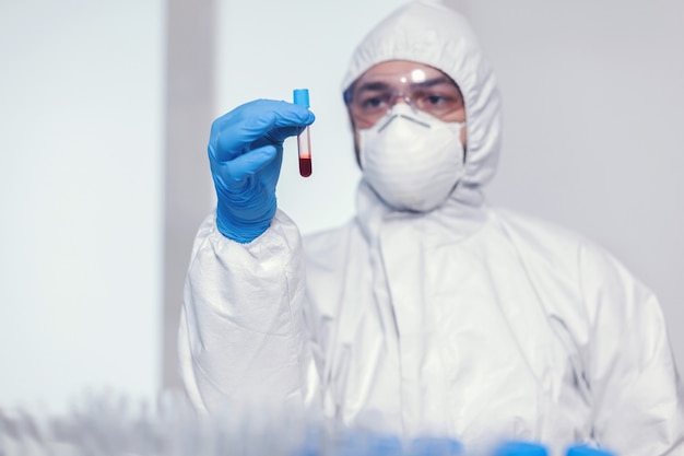 Hombre en traje de mono en laboratorio de microbiología con tubo de ensayo con sangre infectada con coronavirus. Médico que trabaja con diversas bacterias y tejidos, investigación farmacéutica de antibióticos contra