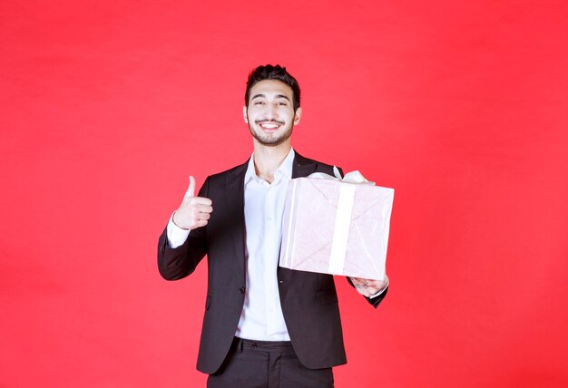 Hombre de traje negro sosteniendo una caja de regalo de color púrpura y mostrando un signo de mano positivo.