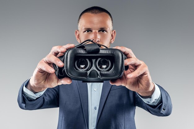 Un hombre con traje que muestra gafas de realidad virtual sobre un fondo gris.