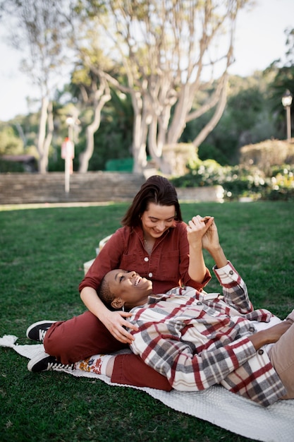 Hombre trans sonriendo y manteniendo la cabeza en el regazo de su novia mientras hace un picnic en el parque