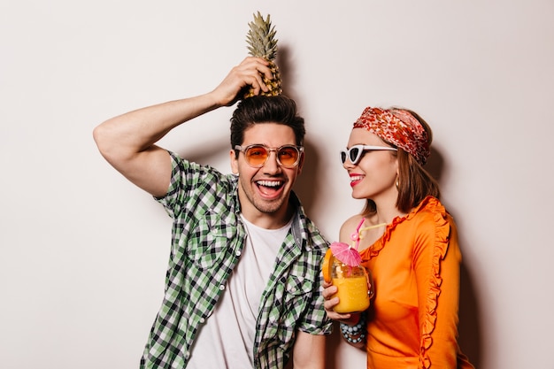 Hombre travieso con camisa verde tiene piña en la cabeza y mira a la cámara con una sonrisa. Dama de blusa naranja y vasos con cóctel.