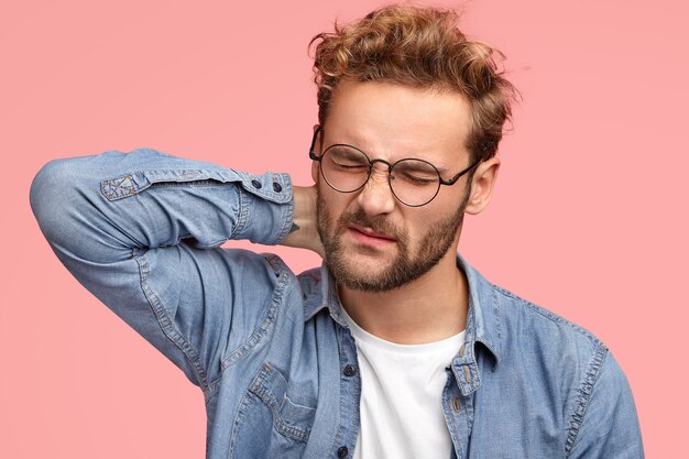 El hombre triste se pone rígido en el cuello, sufre de dolor, tiene un estilo de vida sedentario y trabaja durante mucho tiempo en la computadora, frunce el ceño en señal de insatisfacción, usa anteojos y camisa de mezclilla, se para en el interior