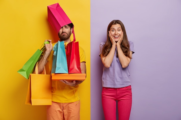 Hombre triste sobrecargado con bolsas de compras, esposa adicta a las compras, dedica tiempo libre durante el fin de semana comprando ropa nueva