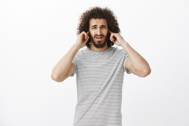 Hombre, tu juego es horrible. Retrato de chico hispano atractivo incómodo disgustado con barba y peinado afro, frunciendo el ceño, cubriendo las orejas con los dedos índices, escuchando ruidos molestos