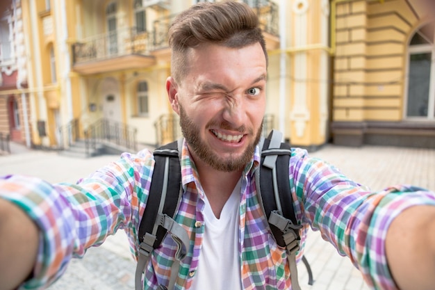 Foto gratuita hombre turista haciendo sonrisa de foto selfie. hombre barbudo en camisa a cuadros con mochila sonriendo para la cámara al aire libre en la calle de la ciudad.