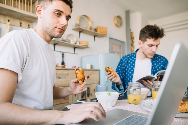 Hombre usando una computadora portátil en la mesa de comedor con su amigo leyendo un libro y desayunando