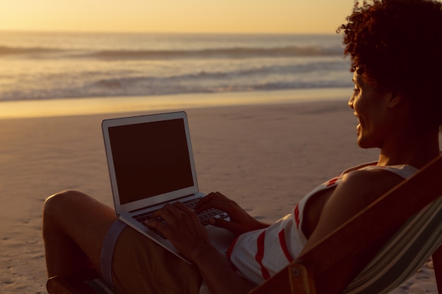Hombre usando laptop mientras se relaja en una silla de playa en la playa