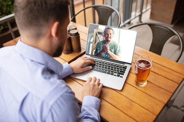 Hombre usando laptop para videollamada mientras bebe una cerveza