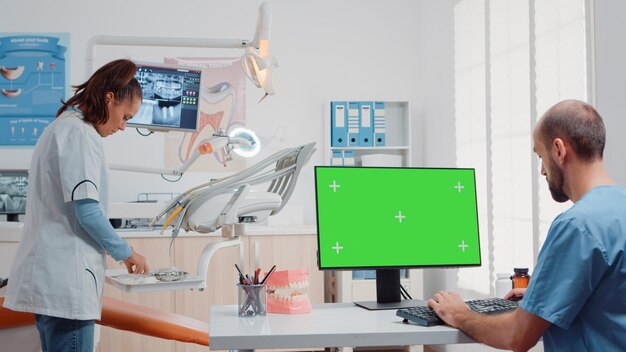 Hombre usando un monitor con pantalla verde horizontal y hablando con el dentista sobre el cuidado bucal en el gabinete. Especialistas que trabajan con herramientas dentales para el cuidado de los dientes y plantilla de maqueta aislada en la computadora