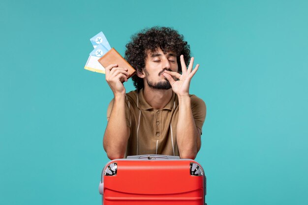 Hombre de vacaciones sosteniendo boletos y apoyado en su bolsa roja sobre azul