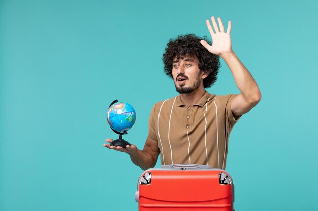 Hombre de vacaciones sosteniendo globo terráqueo con bolsa roja ondeando sobre azul