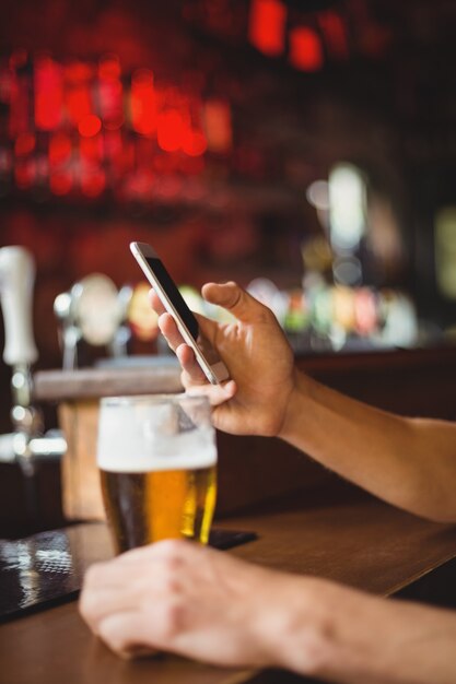 Hombre con vaso de cerveza mediante teléfono móvil en mostrador