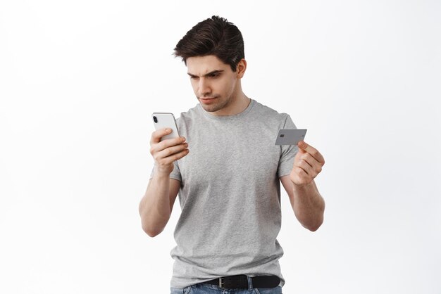El hombre se ve serio en la pantalla del teléfono inteligente mientras realiza compras de pago en línea con la entrega de pedidos de tarjetas de crédito de plástico en Internet de pie sobre fondo blanco.