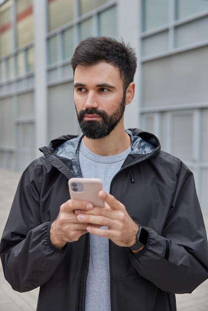 El hombre vestido con anorak negro utiliza el teléfono móvil para chatear, la aplicación de descargas en línea para poses de entrenamiento deportivo al aire libre en borrosa. Estilo de vida moderno