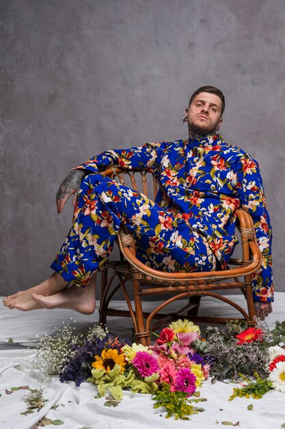 Un hombre en vestido floral sentado en una silla con diferentes flores en el piso