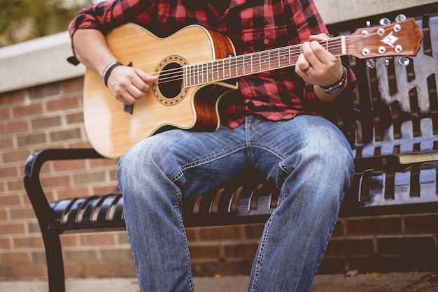 Hombre vestido con una franela roja y negra sentado en un banco tocando la guitarra