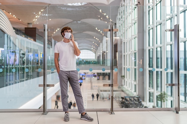 Hombre vestido con mascarilla en el aeropuerto y hablando por teléfono inteligente