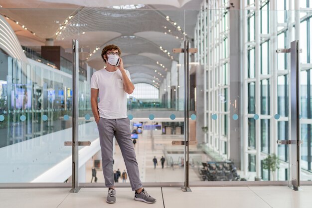 Hombre vestido con mascarilla en el aeropuerto y hablando por teléfono inteligente