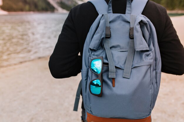 Hombre vestido con un suéter de algodón negro de pie frente al lago con una elegante bolsa de viaje y gafas de sol