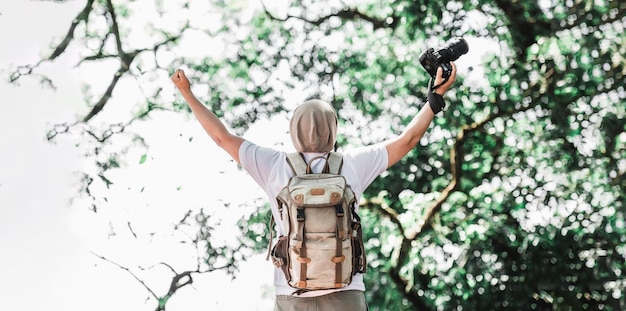 Hombre viajero asiático con mochila sosteniendo una cámara y haciendo un gesto feliz en el bosque con espacio de copia Fotógrafo de viajes Éxito vocacional y concepto de vacaciones