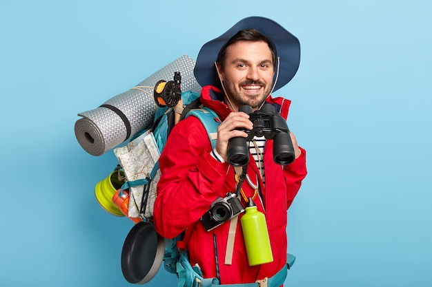 El hombre viajero camina con mochila, lleva las cosas necesarias para el viaje, mira a través de binoculares, tiene una mirada alegre, usa ropa informal