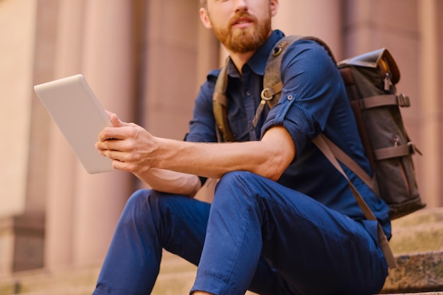 El hombre viajero casual barbudo se sienta en un escalón y usa una tableta.