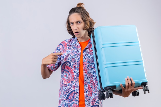 Hombre viajero joven disgustado con maleta apuntando con el dedo hacia él mirando confundido de pie