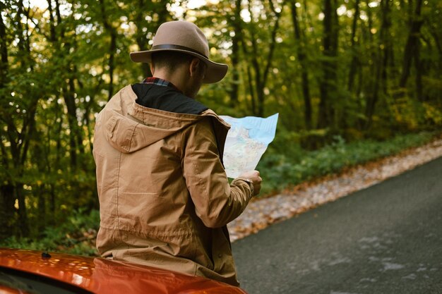 Hombre viajero con sombrero mirando mapa cerca del coche en el bosque de otoño