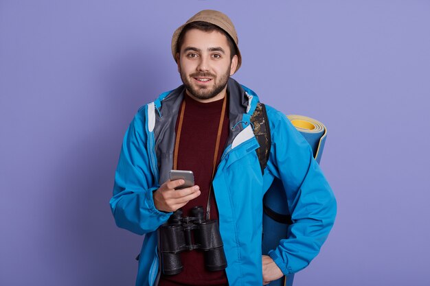 Hombre viajero sonriente con gorra y mochila. Turista que viaja en una escapada de fin de semana, de buen humor, sosteniendo un teléfono inteligente