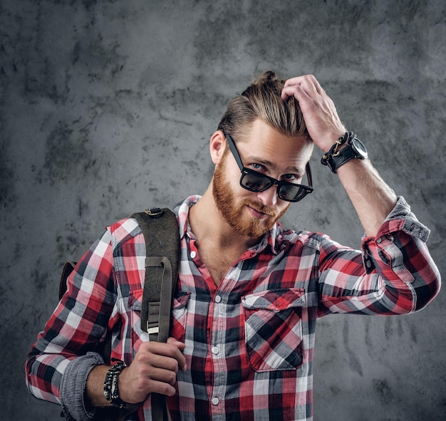 Hombre viajero urbano pelirrojo elegante con gafas de sol sostiene mochila sobre fondo gris en un estudio.