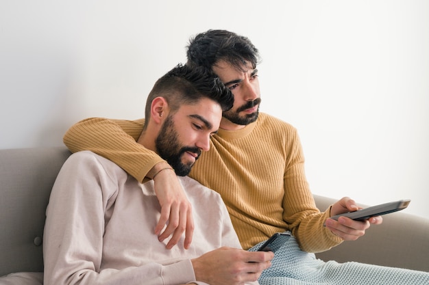 Foto gratuita hombre viendo televisión sentado cerca de su novio usando un teléfono móvil contra una pared blanca