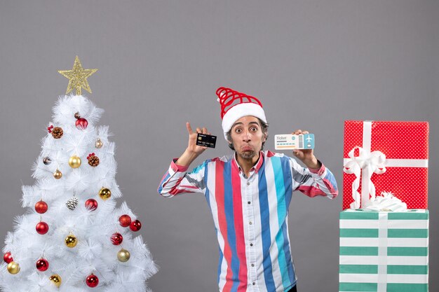 Hombre de vista frontal con miradas indiscretas con tarjeta y boleto de viaje alrededor del árbol de Navidad y regalos