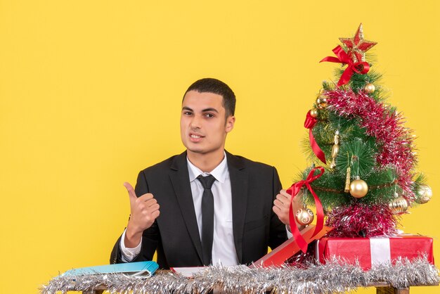 Hombre de vista frontal en traje sentado en la mesa sosteniendo el documento que muestra algo árbol de Navidad y regalos