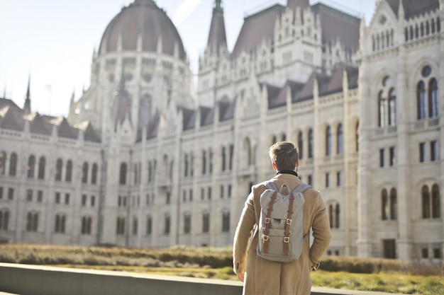 Hombre vistiendo abrigo marrón y mochila cerca del edificio del Parlamento húngaro en Budapest, Hungría