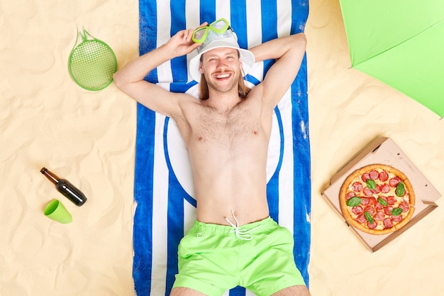 Foto gratuita el hombre yace sobre una toalla a rayas disfruta de un día de verano perezoso en la playa usa sombrero para el sol máscara de snorkel come pizza apetitosa se ha relajado mira felizmente a la cámara toma el sol descansa en un resort tropical